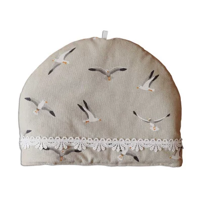 Ein beiger Teewärmer aus Stoff mit Möwenillustrationen und einer weißen Spitzenborte am unteren Rand. Der Teewärmer hat eine kleine Schlaufe an der Oberseite für die Handhabung.