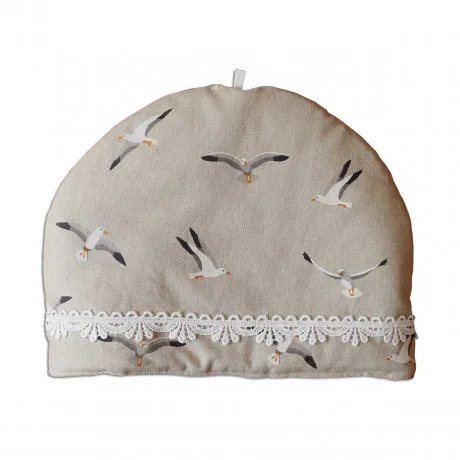 Ein beiger Teewärmer aus Stoff mit Möwenillustrationen und einer weißen Spitzenborte am unteren Rand. Der Teewärmer hat eine kleine Schlaufe an der Oberseite für die Handhabung.