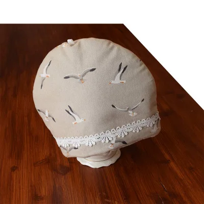 Ein beiger Teewärmer aus Stoff mit einem fliegenden Möwenmuster und weißem Spitzenbesatz am Boden. Er steht auf einer hölzernen Oberfläche, wobei oben und auf der linken Seite ein weißer Hintergrund zu sehen ist.