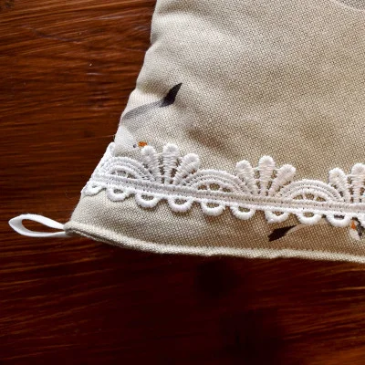 Nahaufnahme eines hellbeigen Stoffes mit feinen dunklen Flecken, der mit einer weißen Spitzenborte und einer kleinen Stoffschleife auf der linken Seite verziert ist. Der Hintergrund ist eine braune Holzoberfläche, die sichtbare Holzmaserungen aufweist.