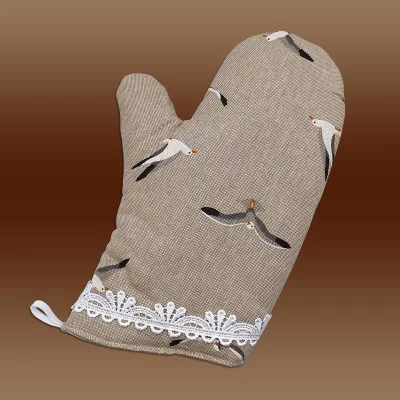 Abgebildet ist ein beigefarbener Topfhandschuh mit einem fliegenden Möwenmuster und weißem Spitzenbesatz an der Manschette. Der Hintergrund ist braun mit Farbverlauf, und an der unteren Ecke des Handschuhs ist eine kleine weiße Aufhängeschlaufe angebracht.