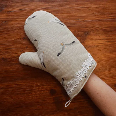 Die linke Hand trägt einen beigen Topfhandschuh mit Möwenmotiven und weißer Spitzenborte am Handgelenk. Der Hintergrund besteht aus einer Holzoberfläche.