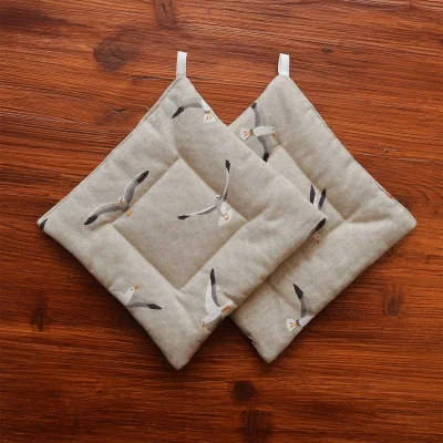 Zwei beigefarbene, quadratische, gesteppte Topflappen mit Möwenmotiv sind auf einer Holzfläche platziert. Jeder hat einen abgesteppten Rand und eine kleine weiße Aufhängeschlaufe in einer Ecke. Das Holz hat eine natürliche, strukturierte Oberfläche.