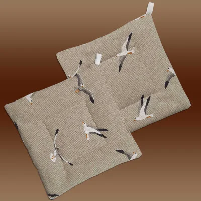 Zwei quadratische beigefarbene Topflappen aus Stoff mit dezenter Webstruktur und einem aufgedruckten Muster aus fliegenden Möwen. Jeder Topflappen hat einen genähten quadratischen Rand und eine weiße Stoffschlaufe zum Aufhängen. Der Hintergrund zeigt einen sanften braunen Farbverlauf.
