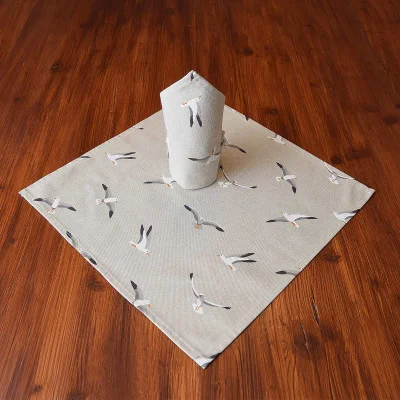 Eine gefaltete Serviette steht aufrecht auf einem passenden quadratischen Tischset; beide sind hellgrau mit einem Muster aus fliegenden Möwen. Das Set aus Serviette und Tischset steht auf einem Holztisch mit sattbrauner Oberfläche.