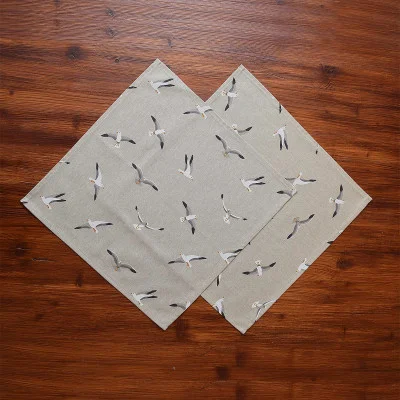 Zwei beigefarbene Stoffservietten mit fliegendem Möwenmuster sind rautenförmig angeordnet, überlappen sich teilweise in der Mitte und liegen auf einer Holzfläche mit deutlicher brauner Maserung.