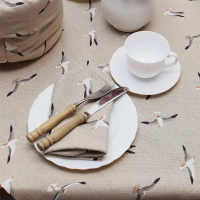 Eine weiße Teetasse und eine Untertasse stehen auf einem beigen Tischtuch mit einem fliegenden Möwenmuster. Daneben stehen ein weißer Teller mit einer gefalteten Serviette sowie eine Gabel und ein Messer mit Holzgriffen.