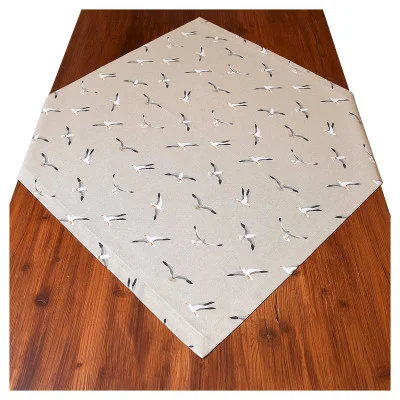 Eine hellbeige Mitteldecke mit einem Muster aus weißen Möwen mit grauen und schwarzen Akzenten ist diagonal auf einem Holztisch angeordnet. Die Holzmaserung des Tisches ist an den Rändern des Läufers sichtbar.