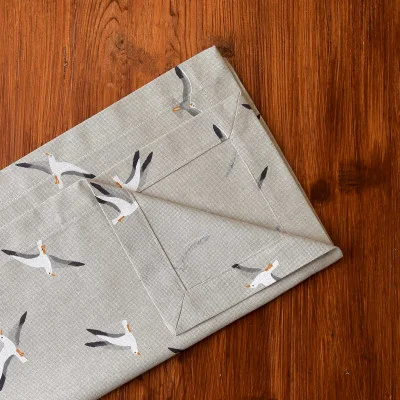 Ein gefaltetes hellgraues Tuch mit einem Muster aus fliegenden Möwen liegt auf einer rotbraunen Holzfläche. Das Tuch weist sichtbare Nähte auf und ist sauber gefaltet, wobei eine Ecke überlappt und das Vogelmuster hervorgehoben wird.