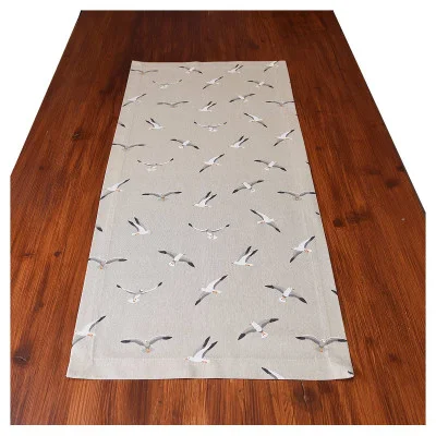 Ein brauner Holztisch mit einem beigen Tischläufer, der ein Muster aus fliegenden Möwen in verschiedenen Positionen zeigt. Der Tischläufer ist mittig auf dem Tisch platziert, und der Hintergrund ist einfarbig weiß.