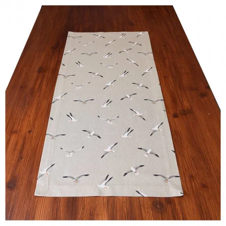 Ein brauner Holztisch mit einem beigen Tischläufer, der ein Muster aus fliegenden Möwen in verschiedenen Positionen zeigt. Der Tischläufer ist mittig auf dem Tisch platziert, und der Hintergrund ist einfarbig weiß.