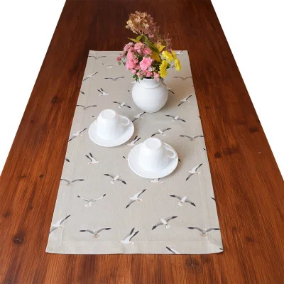 Ein Holztisch ist mit einem beigen Tischläufer bedeckt, der mit einem Möwenmuster verziert ist. Auf dem Tisch stehen eine weiße Vase mit rosa und gelben Blumen und zwei weiße Teetassen auf Untertassen, die neben der Vase aufgestellt sind.