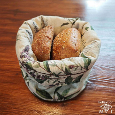 Zwei braune Brötchen liegen in einem Stoffkorb mit Blumenmuster auf einer Holzoberfläche. Der Korb ist mit grünen Blättern und violetten Blumen verziert, was der Präsentation eine rustikale Note verleiht.