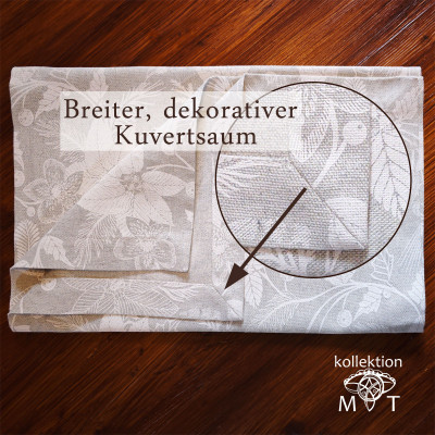 Gefaltete beige Tischdecke mit Blumenmuster auf einem Holztisch. Eine Lupe hebt den breiten, dekorativen Saum mit der Aufschrift „Breiter, dekorativer Kuvertsaum“ hervor. In der unteren rechten Ecke steht der Text „kollektion MT“.