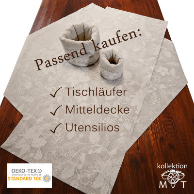 Auf einem Holztisch steht ein Satz grauer Tischwäsche mit Blumenmuster, darunter ein Tischläufer, ein Tafelaufsatz und Utensilienhalter. Der deutsche Text lautet: „Passend kaufen: Tischläufer, Mitteldecke, Utensilios.“ OEKO-TEX- und kollektion MT Logos sind sichtbar.