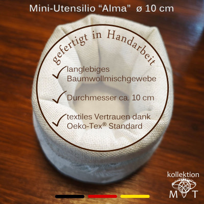 Ein kleiner, handgefertigter Stoffbehälter mit der Aufschrift „Mini-Utensilio ‚Alma‘ ø 10 cm“. Eigenschaften: strapazierfähige Baumwollmischung, 10 cm Höhe, Öko-Tex-Standard-Zertifizierung. Hintergrund ist eine Holzoberfläche mit deutscher Textauflage.