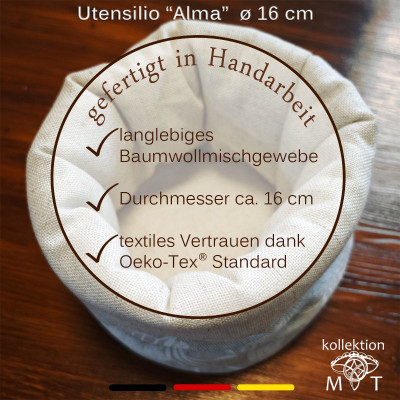 Ein runder, beiger Stoffbehälter mit der Aufschrift „Utensilio ‚Alma‘ Ø 16 cm“ auf einer Holzoberfläche. Der deutsche Text hebt handgefertigte Handwerkskunst, langlebige Baumwollmischung, 16 cm Durchmesser und Öko-Tex-Standard hervor. Das Kollektionslogo in den Farben der deutschen Flagge ist sichtbar.