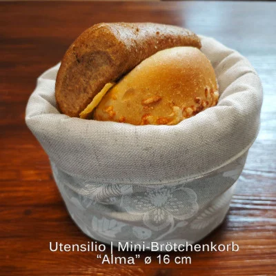 Ein kleiner Brotkorb mit der Aufschrift „Utensilio | Mini-Brötchenkorb 'Alma' ø 16 cm“ hält zwei Brotsorten, eine helle mit Käse und eine dunkle, auf einem Holztisch.