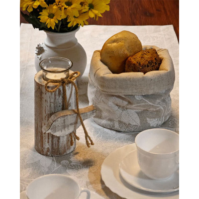 Ein rustikaler Tisch mit einer naturfarbenen Tischdecke, einem dekorativen Kerzenständer mit Schleife, einem Stoffkorb mit zwei Brötchen, weißen Teetassen auf Untertassen und einer Vase mit gelben Blumen.