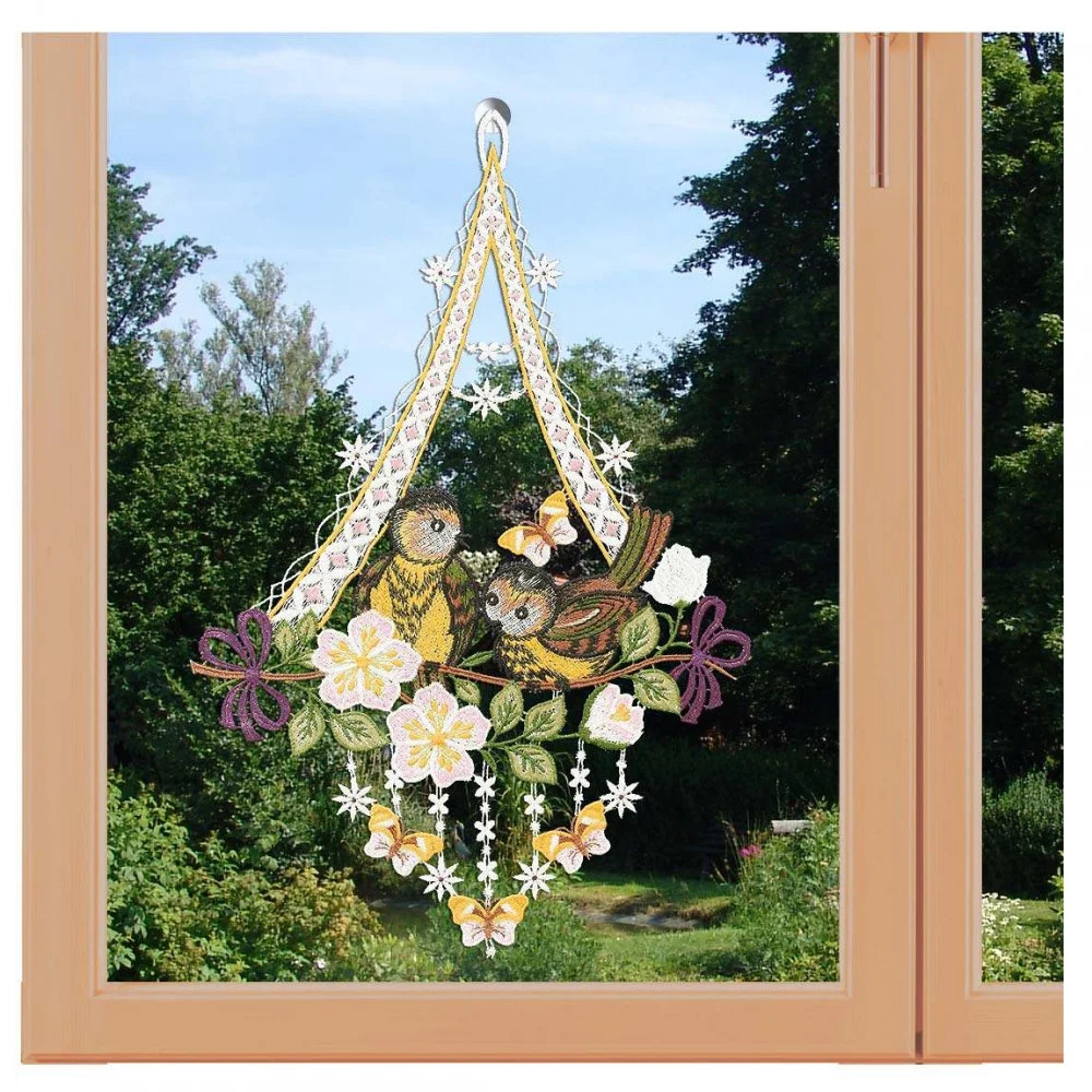 Ein dekoratives Fensterbild mit Vögeln auf einem Ast, Blumen und Schmetterlingen hängt in einem hölzernen Fensterrahmen. Die Szene fängt die Essenz der Vogtländischen Spitze ein, mit üppigem Grün und Bäumen unter einem klaren Himmel dahinter.