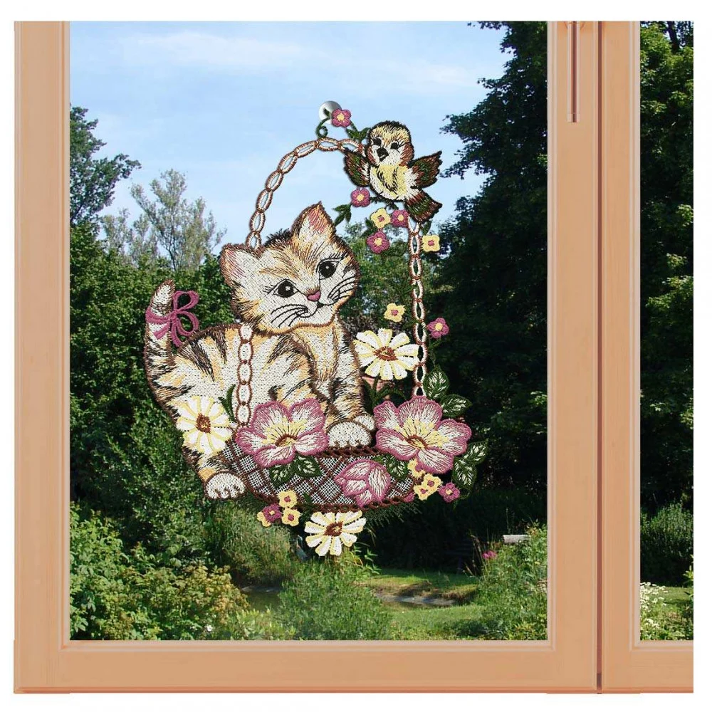 Ein Fensterbild zeigt ein süßes Kätzchen in einem blumengeschmückten Korb, über dem ein kleiner Vogel thront. Der Hintergrund zeigt einen Gartenblick mit üppig grünen Bäumen und Büschen unter klarem Himmel, zart eingerahmt von Vogtländischer Spitze.