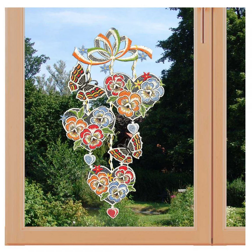 Ein Fenster, geschmückt mit farbenfroher Spitzenstickerei mit Blumen und Schmetterlingen, ergänzt durch einen üppigen Gartenblick. Der Holzrahmen verleiht dem filigranen Design der Vogtländischen Spitze, das als Fensterbild hängt, Wärme und verbindet Eleganz mit der Kunstfertigkeit der Natur.
