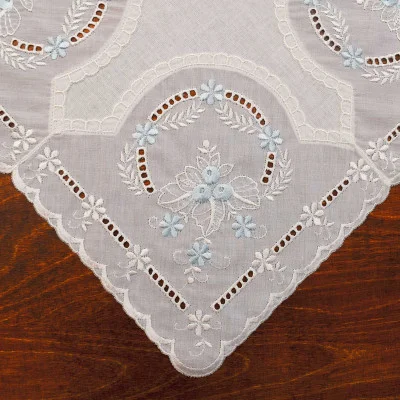 Eine weiße, bestickte Tischdecke mit gewellten Rändern und Blumen- und Blattmustern aus weißem und hellblauem Garn ergibt auf einer Holzoberfläche eine bezaubernde Tischdekoration aus Vogtländischer Spitze.