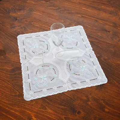 Auf einem Holztisch steht ein klares, leeres Sektglas, platziert auf einem quadratischen Deckchen aus vogtländischer Spitze, das mit hellblauen Blumenmustern verziert ist – ein eleganter Hauch von Tischdekoration.