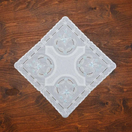 Ein quadratisches, weißes Baumwollbatist-Deckchen aus Vogtländischer Spitze mit hellblauer Blumenstickerei liegt diagonal auf einer Holzoberfläche. Dieses elegante Deckchen besticht durch sein aufwendiges Ausschnittmuster und die gewellten Kanten – perfekt als Tischdekoration.