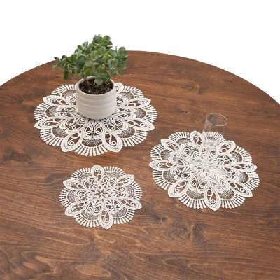 Auf einem runden Holztisch liegen drei runde Deckchen aus Vogtländischer Spitze – jedes ziert eine einzigartige Tischdekoration: eine kleine Sukkulente im Topf, ein Trinkglas und ein leeres Spitzendeckchen.