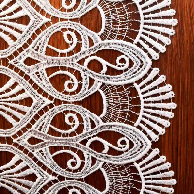 Weißer Spitzenstoff mit komplizierten, wirbelnden Mustern und gewellten Rändern wird auf einer Holzoberfläche präsentiert. Die zarte Vogtländische Spitze ist ideal für eine elegante Tischdekoration geeignet.
