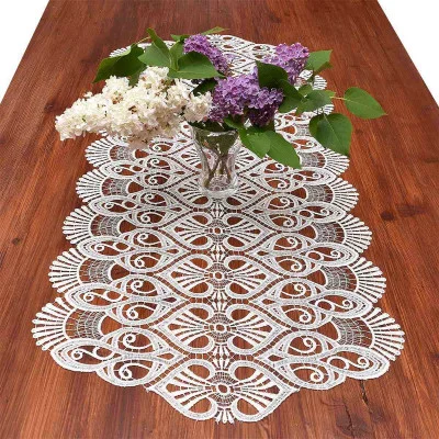 Eine Glasvase mit lila und weißen Flieder steht auf einem Holztisch mit einem kunstvollen vogtländischen Spitzen-Tischläufer Amara, der ein aufwendiges Muster für eine elegante Tischdekoration aufweist.