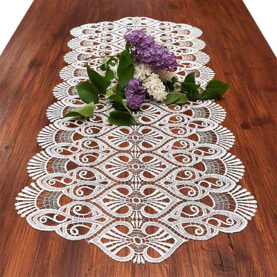 Auf einem Holztisch liegt ein dekorativer Tischläufer Amara aus vogtländischer Spitze und ein kleiner Strauß violetter und weißer Blumen mit grünen Blättern als Tafelaufsatz, wodurch eine elegante Tischdekoration entsteht.