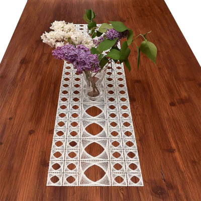 Eine klare Glasvase mit weißem und violettem Flieder steht auf einem Holztisch, in der Mitte ein weißer Tischläufer aus Spitze mit geometrischem Muster.