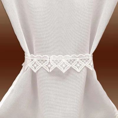 Weißer, transparenter Vorhang, in der Mitte mit einem dekorativen Spitzenraffhalter mit Rauten- und Muschelmuster gerafft, vor braunem Hintergrund.