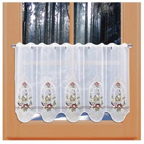 Ein kurzer, dekorativer weißer Vorhang aus Vogtländischer Spitze mit gestickten Vögeln, Äpfeln und Kirchenfenstermotiven hängt an einem Holzrahmen. Draußen sorgt ein verschneiter, dichter Wald für die perfekte Adventsfenster-Stimmung.