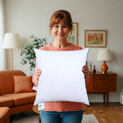 Eine lächelnde Frau mit roten Haaren und einem rosa Pullover steht in einem gemütlichen Wohnzimmer und hält ein weißes quadratisches Kissen in die Kamera. Im Hintergrund sind Retro-Möbel, Pflanzen und gerahmte Kunstwerke zu sehen.