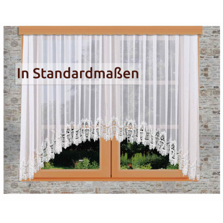 Ein Fenster mit weißen Spitzenvorhängen mit Wellenschliff und Blumenmuster in einem Holzrahmen vor einer grauen Steinwand. Der Text "In Standardmaßen" erscheint auf der linken Seite.