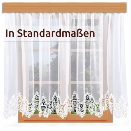 Weiße Spitzenvorhänge mit einem verschnörkelten Blumenmuster hängen an einer Holzstange vor einem Fenster. Ein Etikett mit den Worten "In Standardmaßen" befindet sich oben links auf dem Bild.
