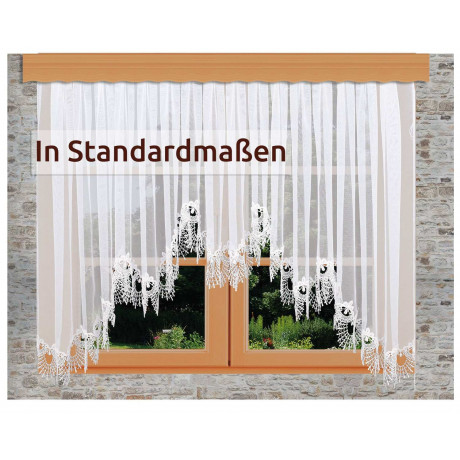 Ein weißer Spitzenvorhang mit floralen Mustern hängt an einer Holzstange über einem Doppelfenster mit Blick ins Grüne. Auf der linken Seite des Bildes steht der Schriftzug "In Standardmaßen".