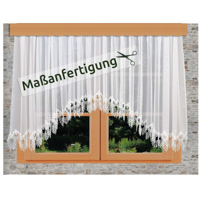 Weißer Spitzenvorhang mit floralen Mustern, der an einem Holzfenster in einer Steinwand hängt. Ein Schild mit Scherensymbol und dem deutschen Text "Maßanfertigung" ist über den Vorhang gelegt.