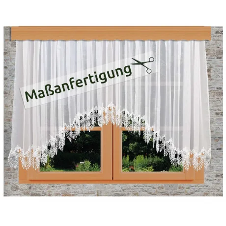 Weißer Spitzenvorhang mit floralen Mustern, der an einem Holzfenster in einer Steinwand hängt. Ein Schild mit Scherensymbol und dem deutschen Text "Maßanfertigung" ist über den Vorhang gelegt.