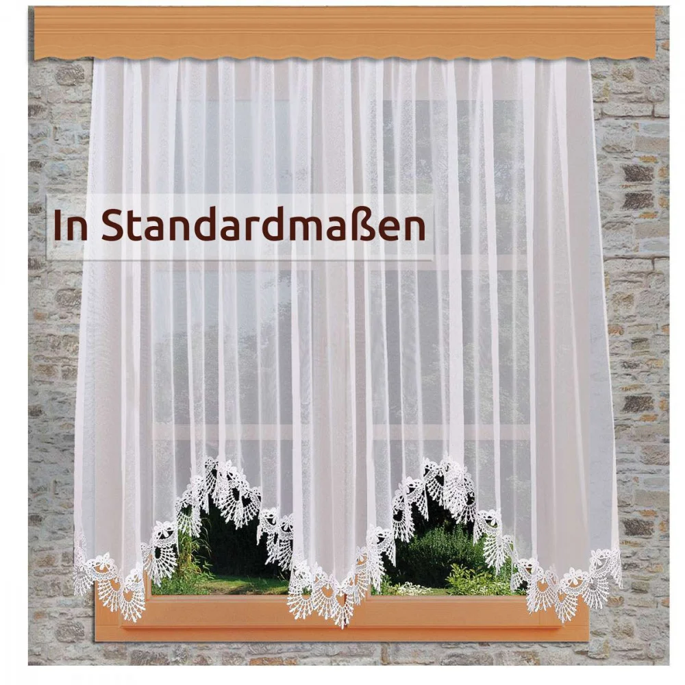 Ein Fenster mit durchsichtigen weißen Spitzenvorhängen, die eine florale Kante aufweisen. Draußen sind grünes Gras und Pflanzen zu sehen. Der Text auf dem Vorhang lautet: "In Standardmaßen.