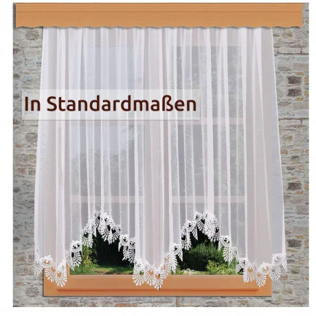 Ein Fenster mit durchsichtigen weißen Spitzenvorhängen, die eine florale Kante aufweisen. Draußen sind grünes Gras und Pflanzen zu sehen. Der Text auf dem Vorhang lautet: "In Standardmaßen.