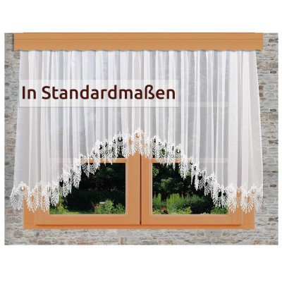 Ein Fenster mit durchsichtigen weißen Spitzenvorhängen, die eine florale Kante aufweisen. Draußen sind grünes Gras und Pflanzen zu sehen. Der Text auf dem Vorhang lautet: "In Standardmaßen.