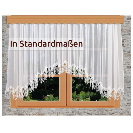 Ein Fenster mit durchsichtigen weißen Spitzenvorhängen, die eine florale Kante aufweisen. Draußen sind grünes Gras und Pflanzen zu sehen. Der Text auf dem Vorhang lautet: "In Standardmaßen.
