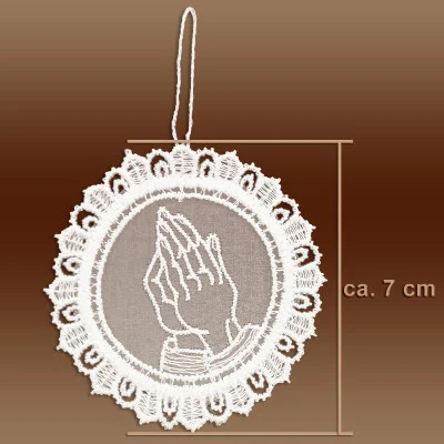 Ein kreisförmiges weißes Spitzenornament aus Plauener Spitze mit betenden Händen in der Mitte, Spitzenbordüren und einer Aufhängeschlaufe. Perfekt als Adventsbaumbehang, er misst ca. 7 cm im Durchmesser. Der Hintergrund ist braun.