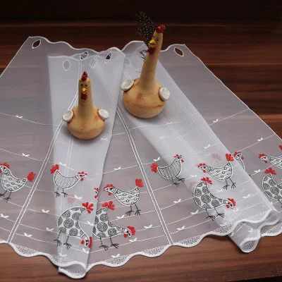 Ein durchsichtiger weißer Vorhang mit gestickten schwarz-weißen Hühnern und roten Akzenten liegt auf einer Holzfläche. Zwei hölzerne Hühnerfiguren mit aufgemalten Gesichtszügen und Federschwänzen sind oben auf dem Vorhang platziert.