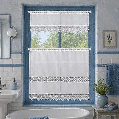 Ein Badezimmer hat ein weißes Waschbecken und blaue Akzente. Natürliches Licht fällt durch ein Fenster mit weißen Spitzenvorhängen und blauer Bordüre ein. Die Wände sind blau und weiß gefliest, und auf einem kleinen Tisch neben der Wanne stehen ein blaues Handtuch, ein gerahmtes Blumenkunstwerk und 