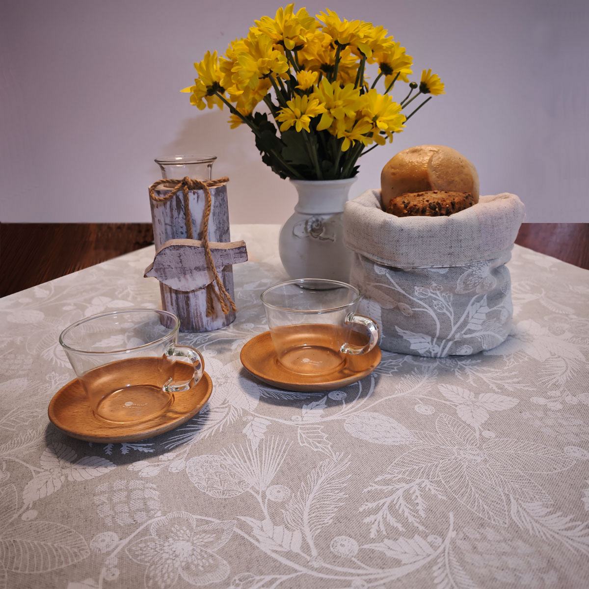 Eine gemütliche Tischdekoration mit zwei Glastassen auf Holzuntertassen, einer Vase mit gelben Blumen, einer rustikalen Kerze und einem mit einem Stoff bedeckten Korb mit Brot, alles auf einer Tischdecke mit hellem Muster.